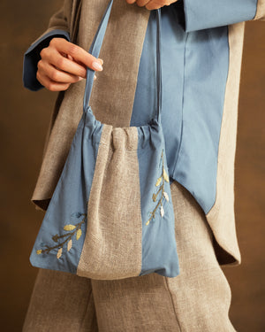 A close-up of a person wearing a blue and beige hand-embroidered drawstring shoulder bag, styled with a matching blue top and beige shirt against a warm brown background.