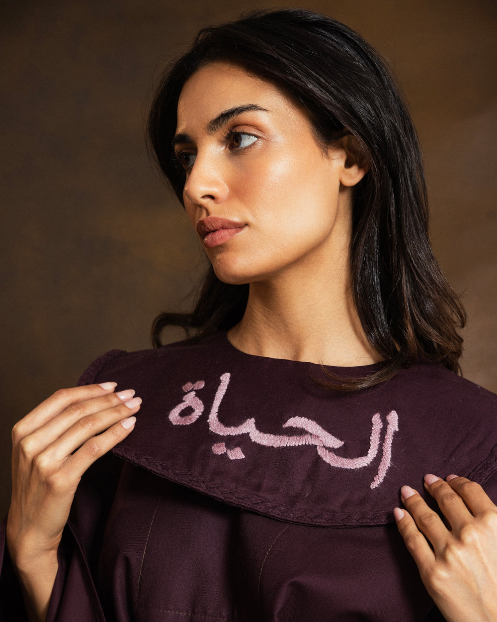 A woman wearing a deep plum purple blouse with a wide collar featuring white Arabic calligraphy, set against a dark brown studio background.