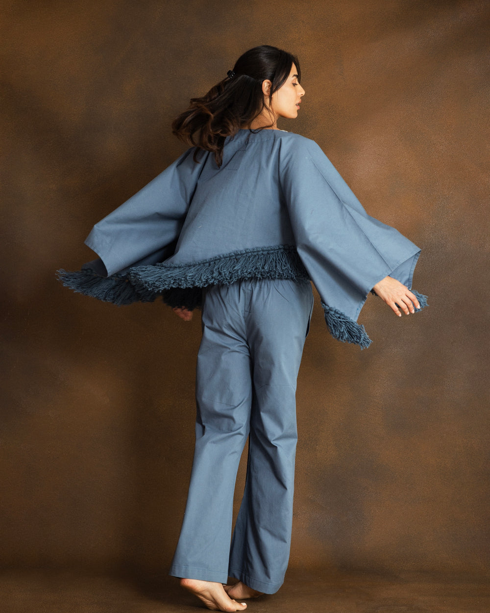 A woman modeling a dusty blue two-piece set consisting of a fringed poncho-style top with silver embellishments and ahnd-embroidered floral motifs and matching wide-leg pants, posed against a textured brown studio backdrop.