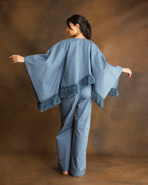 A woman stands against a warm brown studio backdrop, wearing a loose dusty blue poncho-style top with fringe trim and matching wide-leg pants, shown from the back.