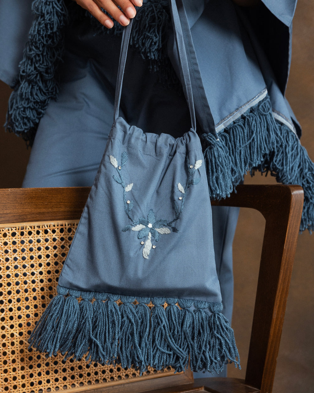 A dusty blue fringed drawstring bag with white floral hand-embroidery, held by a person wearing a matching blue fringed cardigan and trousers, sitting on a wooden cane chair.
