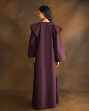 A woman wearing a long, flowing mauve-purple kaftan with wide draped shoulders, shown from the back against a textured brown studio backdrop.