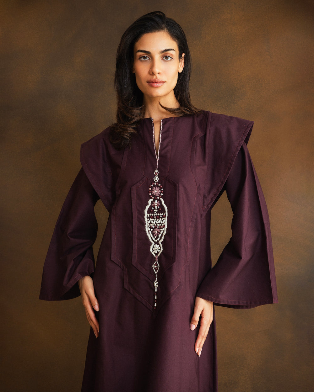 A woman modeling a long, flowing dark plum purple kaftan dress with wide sleeves and a silver-toned statement necklace, standing against a textured brown studio backdrop.