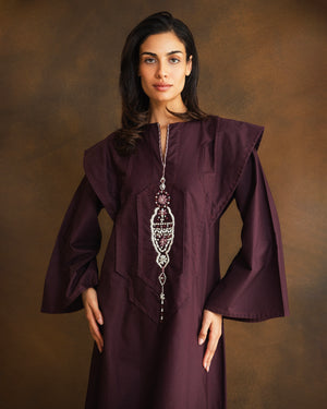 A woman modeling a deep plum purple traditional long robe with wide collar-like shoulder panels and a long ornate silver beaded necklace, set against a warm brown studio backdrop.