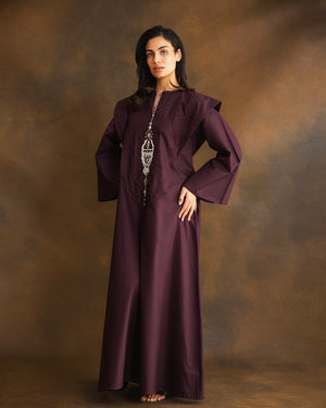 A woman modeling a long, flowing dark plum purple kaftan dress with wide sleeves and a silver-toned statement necklace, standing against a textured brown studio backdrop.
