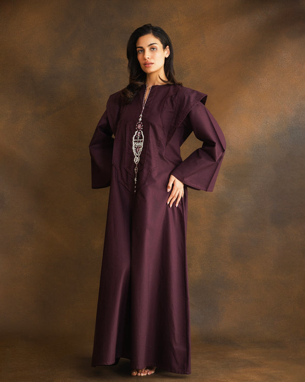 A woman modeling a long, flowing dark plum purple kaftan dress with wide sleeves and a silver-toned statement necklace, standing against a textured brown studio backdrop.