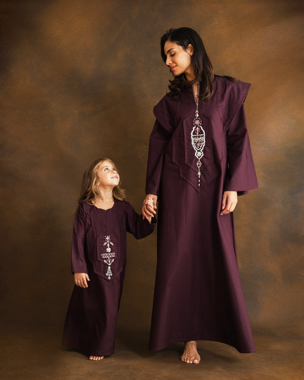 A young girl with long light brown hair stands against a mottled brown studio backdrop, wearing a long burgundy kaftan with white embroidered details on the chest.