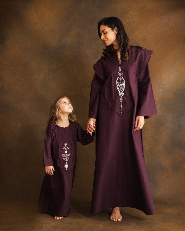 A woman and a young girl wearing matching deep plum-purple traditional long kaftans with silver embroidery, standing against a textured brown studio backdrop.
