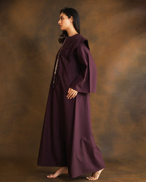 A woman stands in profile wearing a long, flowing dark plum kaftan against a mottled brown studio backdrop.