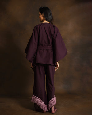 A woman shown from the back wearing a deep plum purple two-piece outfit with wide draped sleeves, a matching waist tie, and fringed hems, set against a dark brown textured studio backdrop.