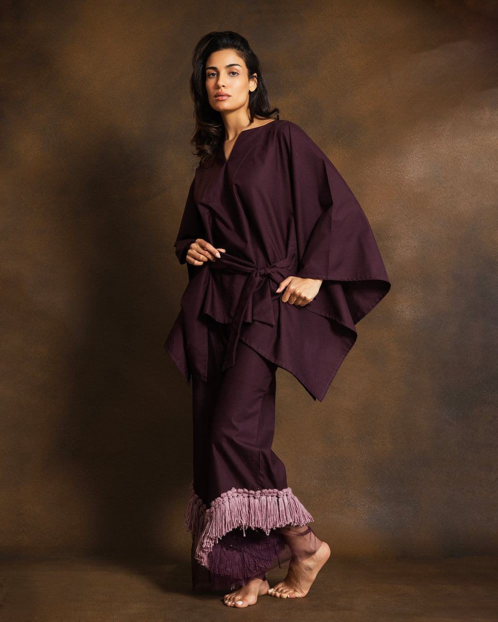 A woman modeling a deep plum purple two-piece outfit consisting of a loose, draped long-sleeve top and matching wide-leg pants with a fringed hem, set against a textured brown studio backdrop.