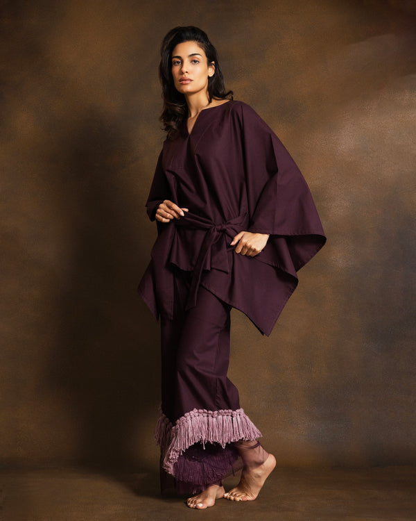 A woman modeling a deep plum purple two-piece outfit consisting of a draped wrap top with a tie belt and matching wide-leg pants featuring a ruffled fringe hem, set against a textured brown studio backdrop.
