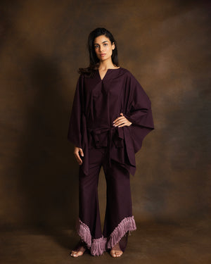 A woman modeling a deep plum purple two-piece outfit consisting of a loose, draped long-sleeve top and matching wide-leg pants with a fringed hem, set against a textured brown studio backdrop.