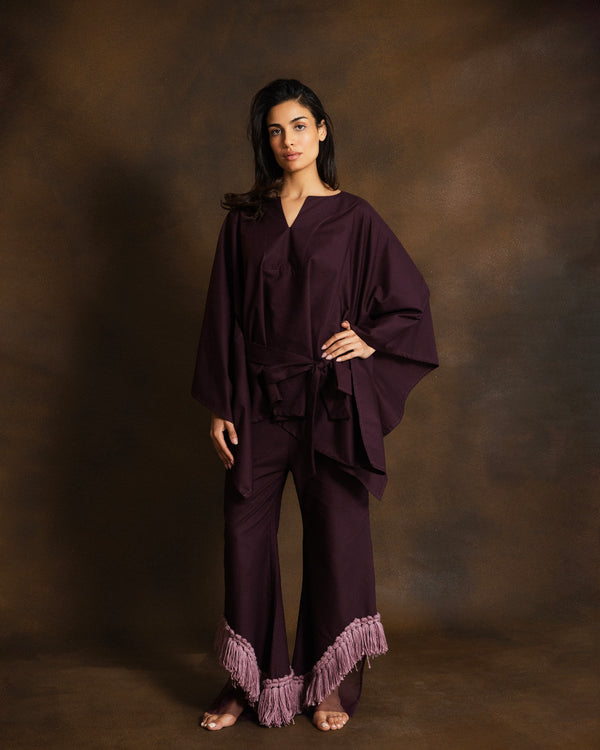 A woman modeling a deep plum purple two-piece outfit consisting of a loose, draped long-sleeve top and matching wide-leg pants with a fringed hem, set against a textured brown studio backdrop.