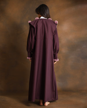 A woman stands facing away from the camera, wearing a long, dark plum-colored dress with pink ruffled trim at the shoulders and cuffs, set against a textured brown studio backdrop.