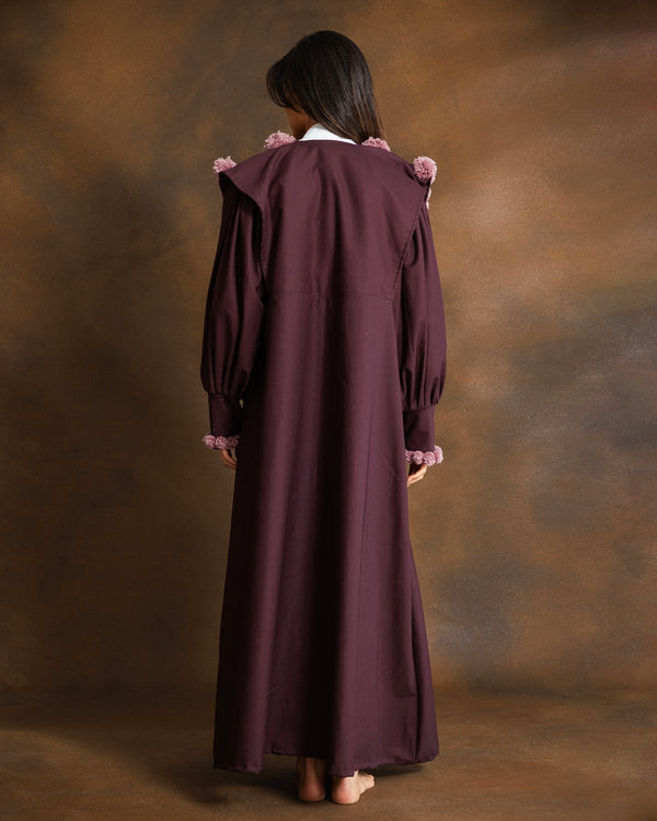 A woman stands facing away from the camera, wearing a long, dark plum-colored dress with pink ruffled trim at the shoulders and cuffs, set against a textured brown studio backdrop.