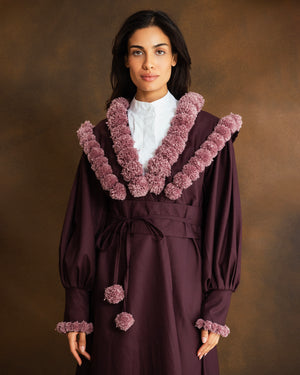 A woman wearing a long burgundy kaftan with oversized dusty mauve pom-pom trim around the collar and cuffs, over a white collared shirt, against a warm brown studio background.
