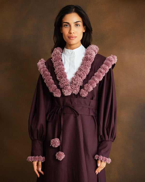 A woman wearing a long burgundy kaftan with oversized dusty mauve pom-pom trim around the collar and cuffs, over a white collared shirt, against a warm brown studio background.