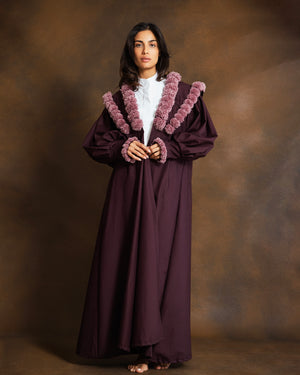 A woman stands against a warm brown studio backdrop wearing a long dark plum robe with voluminous sleeves and a plush mauve-pink textured collar and cuffs over a white shirt.