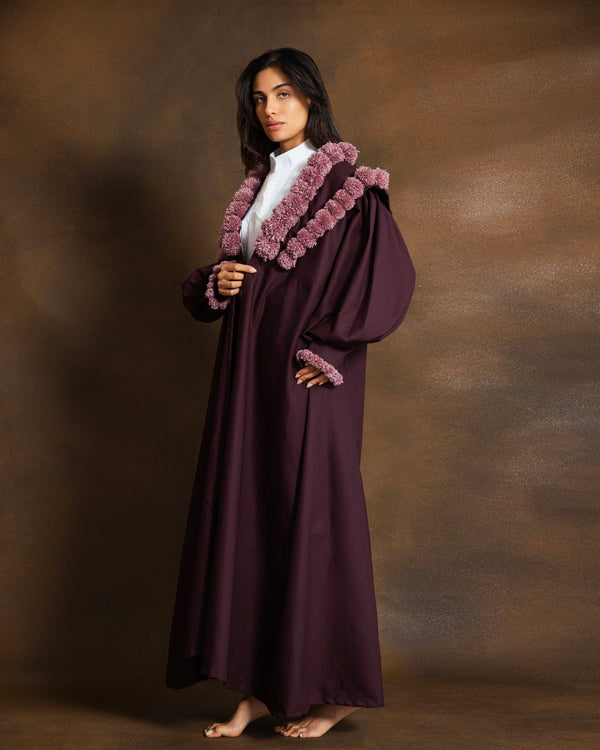 A woman modeling a long, flowing dark plum abaya with a contrasting dusty rose faux-fur collar and cuffs, worn over a white inner top, set against a textured brown studio backdrop.