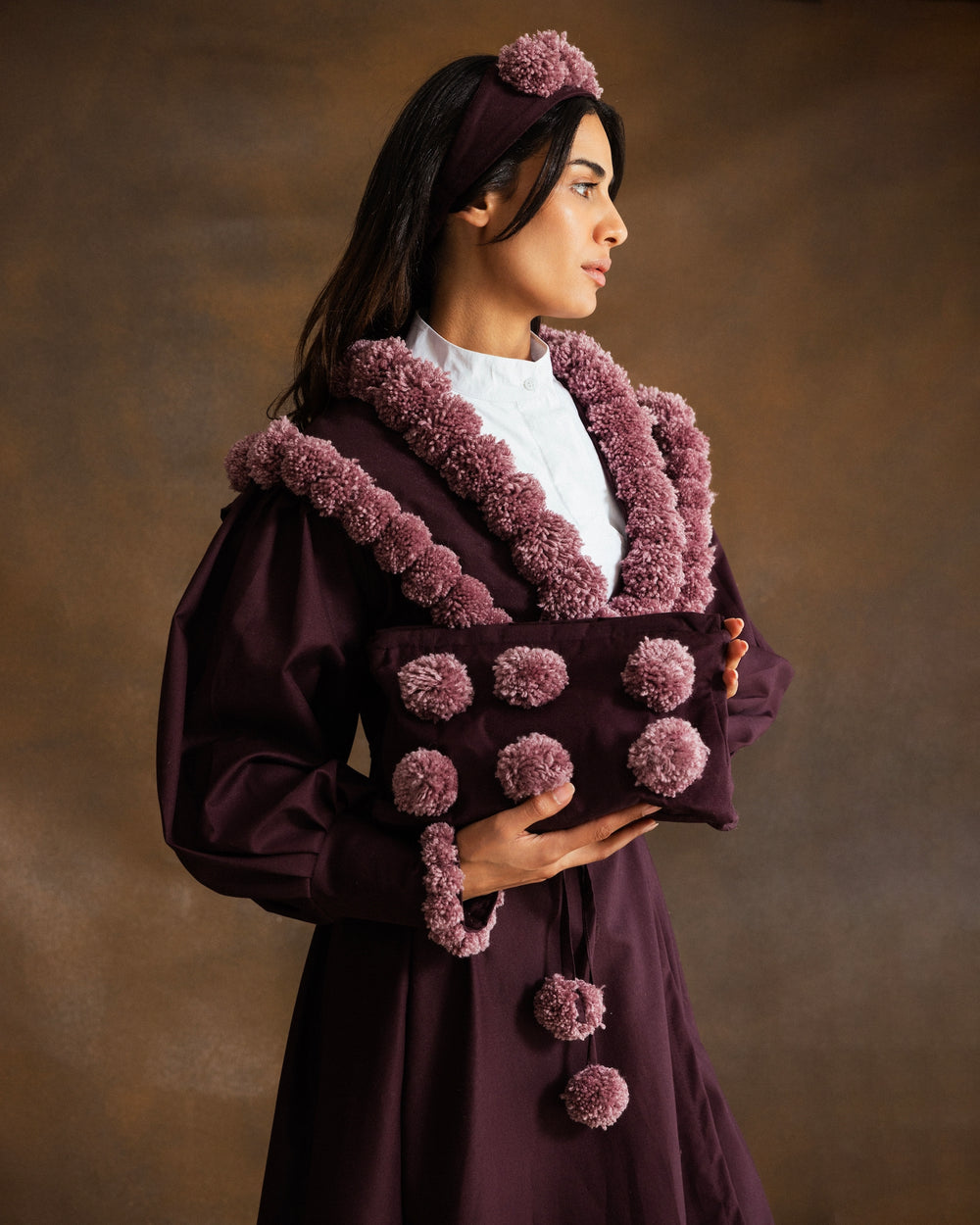 A woman wearing a deep plum purple traditional-style coat with dusty mauve pom-pom trim and a matching headband, holding a matching clutch bag against a warm brown studio background.
