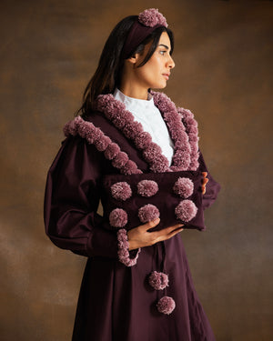 A woman wearing a deep plum purple traditional-style coat with dusty mauve pom-pom trim and a matching headband, holding a matching clutch bag against a warm brown studio background.