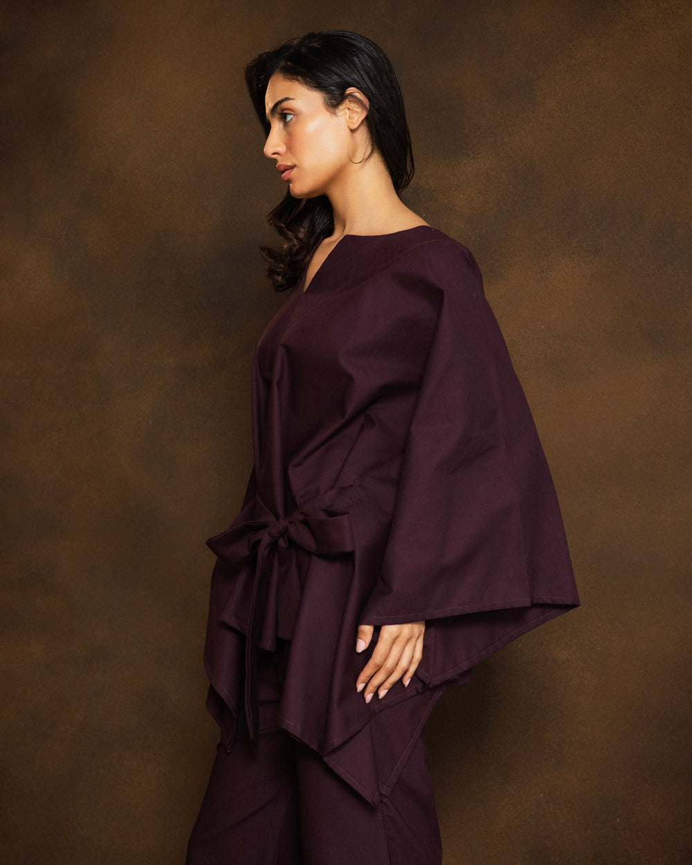 A woman modeling a deep plum purple two-piece outfit consisting of a loose, draped long-sleeve top and matching wide-leg pants with a fringed hem, set against a textured brown studio backdrop.