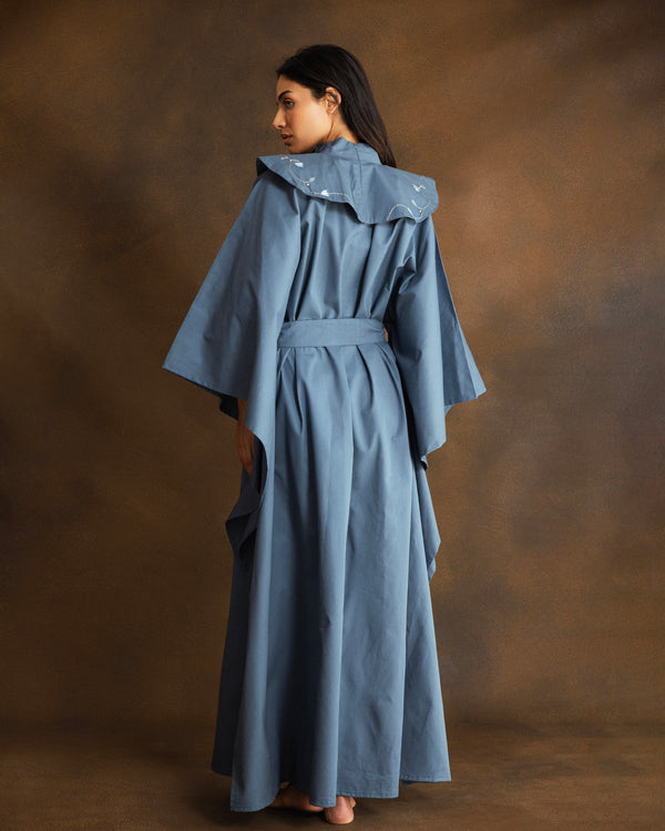 A woman wearing a long, wide-sleeved blue caftan with a matching waist belt, photographed from the back against a warm brown studio backdrop.