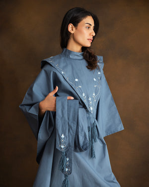A woman wearing a slate blue kaftan with white floral embroidery and tassel details, posed against a warm brown studio backdrop.
