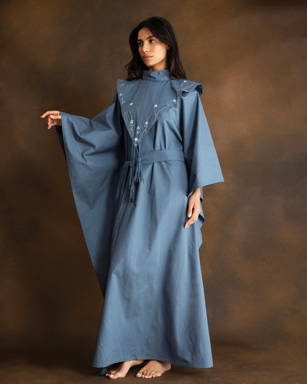 A woman modeling a dusty blue kaftan dress with wide sleeves, a high neck, and silver bead and thread hand-embroidery on the chest, posed against a textured brown studio backdrop.
