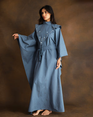 A woman modeling a dusty blue kaftan dress with wide sleeves, a high neck, and silver bead and thread hand-embroidery on the chest, posed against a textured brown studio backdrop.