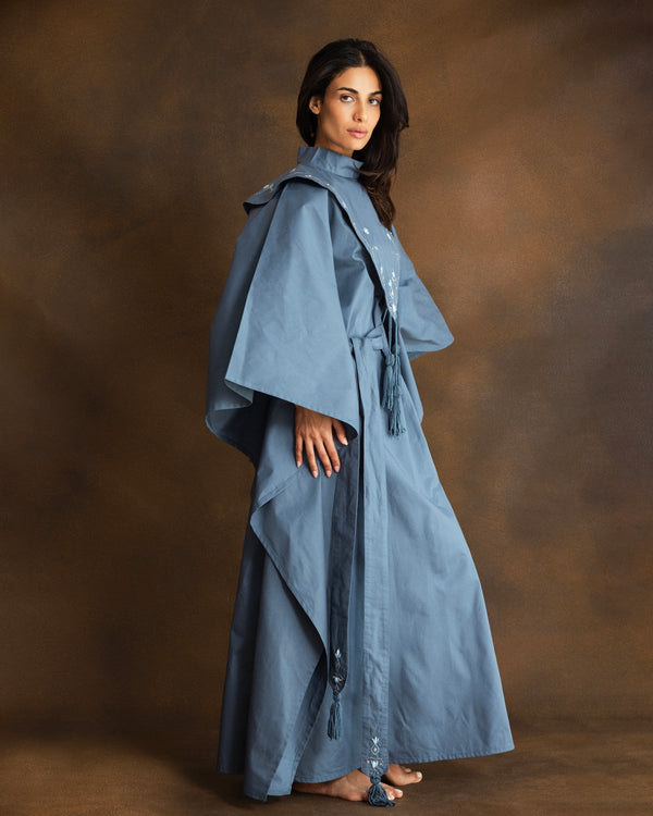 A woman modeling a long, flowing blue caftan with wide draped sleeves and a handmade tassel tie belt, posed against a textured brown studio backdrop.