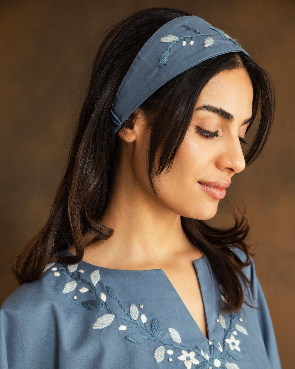 A woman wearing a dusty blue fabric headband with white floral embroidery, paired with a matching blue embroidered top, set against a warm brown background.