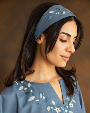 A woman wearing a dusty blue fabric headband with white floral embroidery, paired with a matching blue embroidered top, set against a warm brown background.