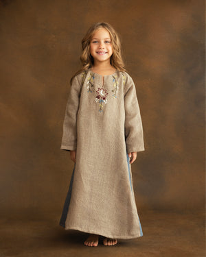Young girl wearing a beige raffia linen kaftan featuring hand-embroidered floral motifs, a crescent moon inspired by Ramadan nights, and dusty blue side panels.