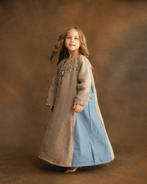 Side view of a young girl wearing a beige raffia linen kaftan with contrasting dusty blue gabardine panels and delicate floral embroidery.