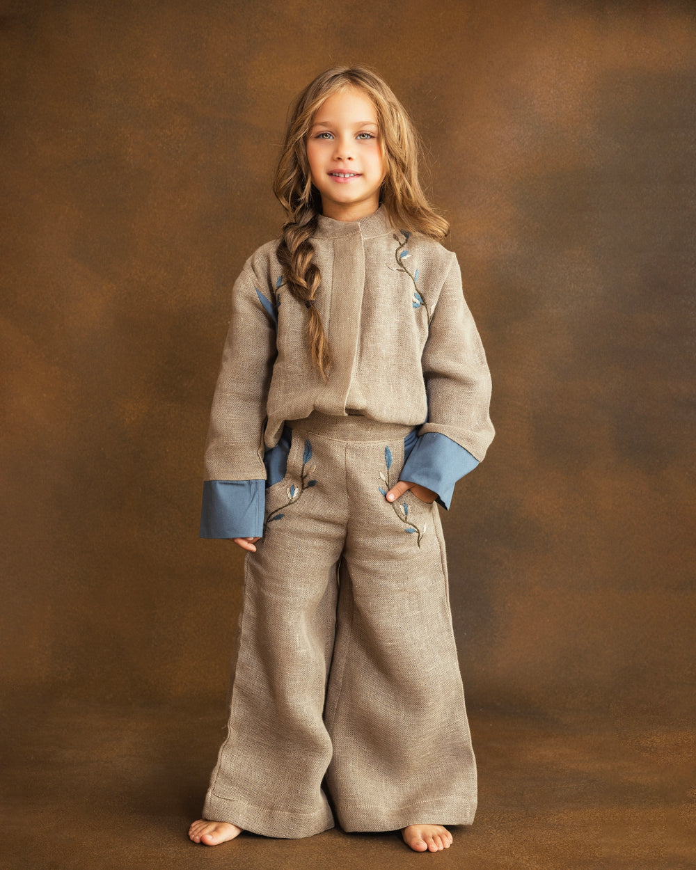 A young girl with long light brown hair stands against a textured brown studio backdrop, wearing a matching beige textured two-piece outfit with blue cuffs and hand-embroidered floral details.