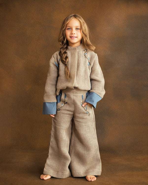 A young girl with long light brown hair stands against a textured brown studio backdrop, wearing a matching beige textured two-piece outfit with blue cuffs and hand-embroidered floral details.