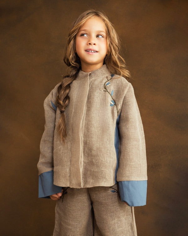 A young girl with a long braided ponytail wearing a beige linen-look jacket with blue contrast panels  with hand-embroidered floral stem motifs and matching beige pants, posed against a textured brown studio backdrop.