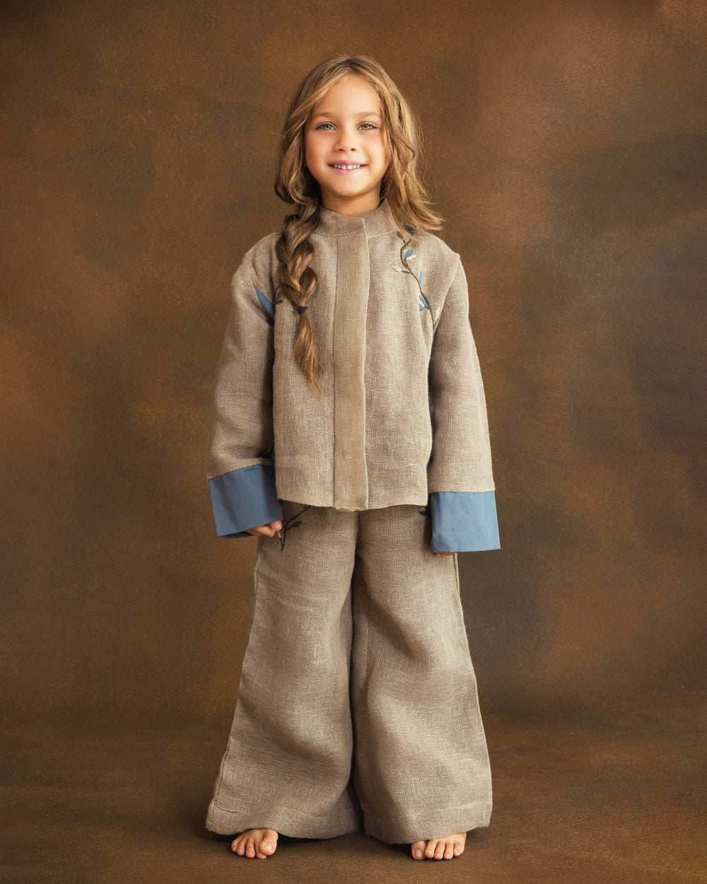 A young girl with long light brown hair stands against a textured brown studio backdrop, wearing a matching beige textured two-piece outfit with contrasting dusty blue cuffs and small blue embroidered details on the chest.