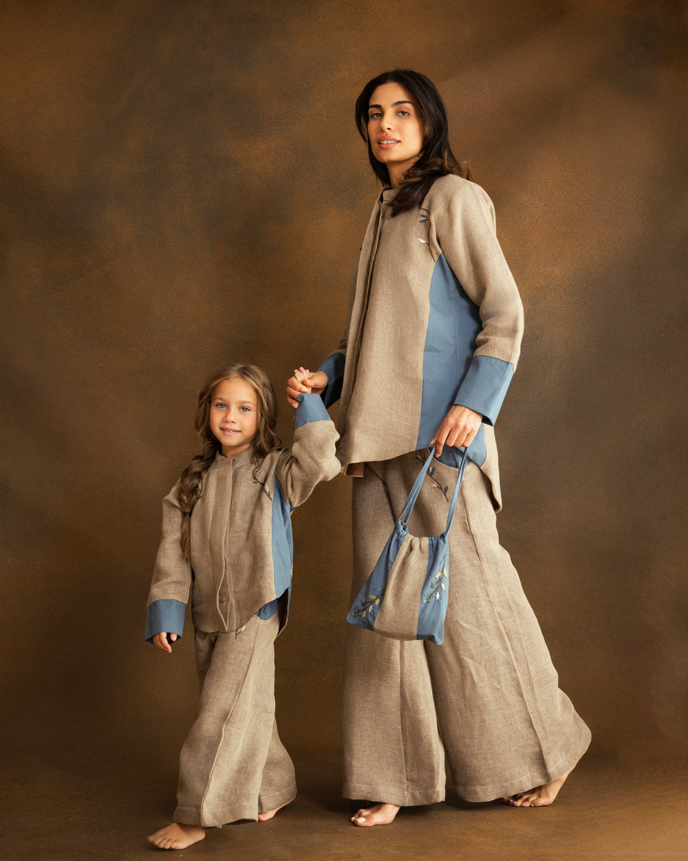 A young girl with long light brown hair stands against a textured brown studio backdrop, wearing a matching beige textured two-piece outfit with contrasting dusty blue cuffs and small blue embroidered details on the chest.