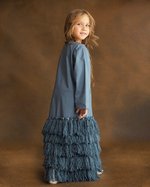 A young girl with long light brown hair stands in profile against a warm brown studio backdrop, wearing a long-sleeve dusty blue dress with a tiered fringe skirt.