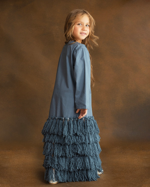A young girl with long light brown hair stands in profile against a warm brown studio backdrop, wearing a long-sleeve dusty blue dress with a tiered fringe skirt.