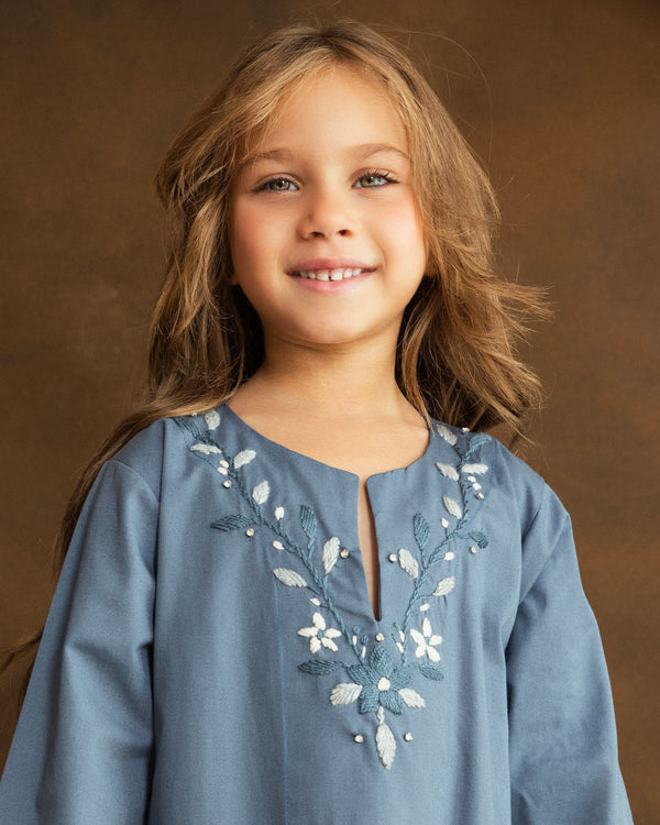 Smiling young girl wearing a dusty blue gabardine kaftan by YMZ Cairo, highlighting delicate blue and ivory floral embroidery and crystal embellishments around the neckline.