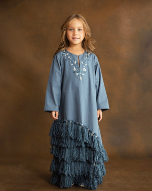 Girl wearing a dusty blue gabardine kaftan by YMZ Cairo, featuring hand-embroidered blue and ivory floral motifs, crystal embellishments, and layered handmade tassels along the asymmetrical hem.