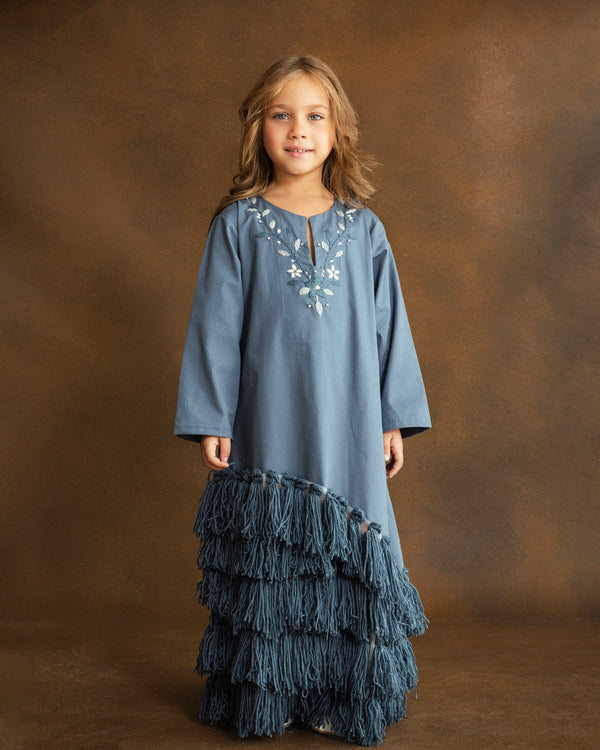Girl wearing a dusty blue gabardine kaftan by YMZ Cairo, featuring hand-embroidered blue and ivory floral motifs, crystal embellishments, and layered handmade tassels along the asymmetrical hem.