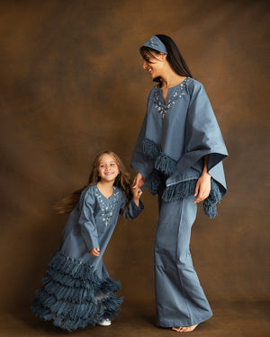 A woman and a young girl wearing matching dusty blue fringed poncho-style dresses and pants sets, standing barefoot against a warm brown studio backdrop. The woman wears a headband and the girl wears shoes, and both are smiling while holding hands.