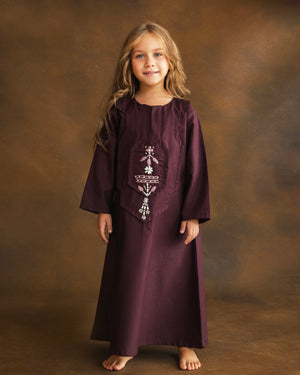 A young girl with long light brown hair stands against a mottled brown studio backdrop, wearing a long burgundy kaftan with white embroidered details on the chest.