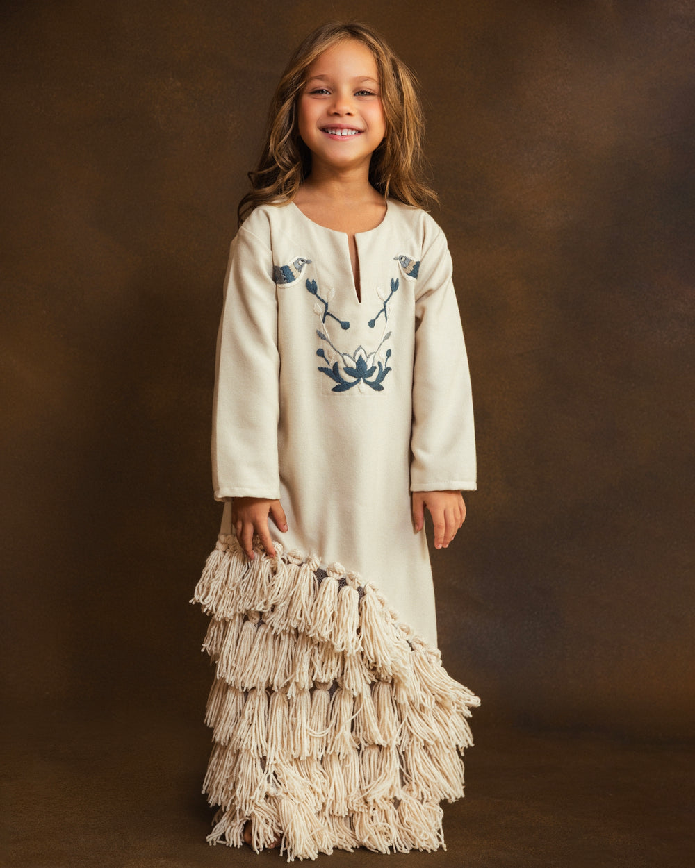 A young girl stands against a textured brown studio backdrop wearing a cream-colored long-sleeve dress with blue floral embroidery on the chest and a tiered handmade tassel hem.