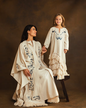Mother-daughter wearing matching cream-colored garments with blue floral embroidery and fringe details, posed against a warm brown studio backdrop.